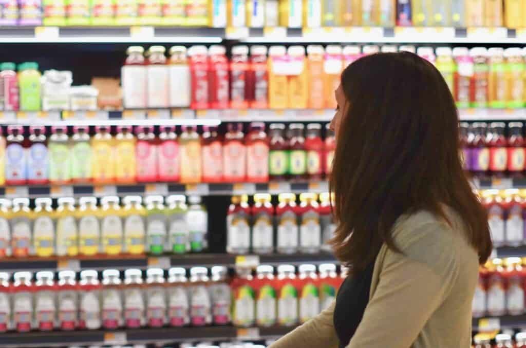 A person with long brown hair stands in a grocery store aisle, looking at shelves filled with various colorful bottled drinks and juices.