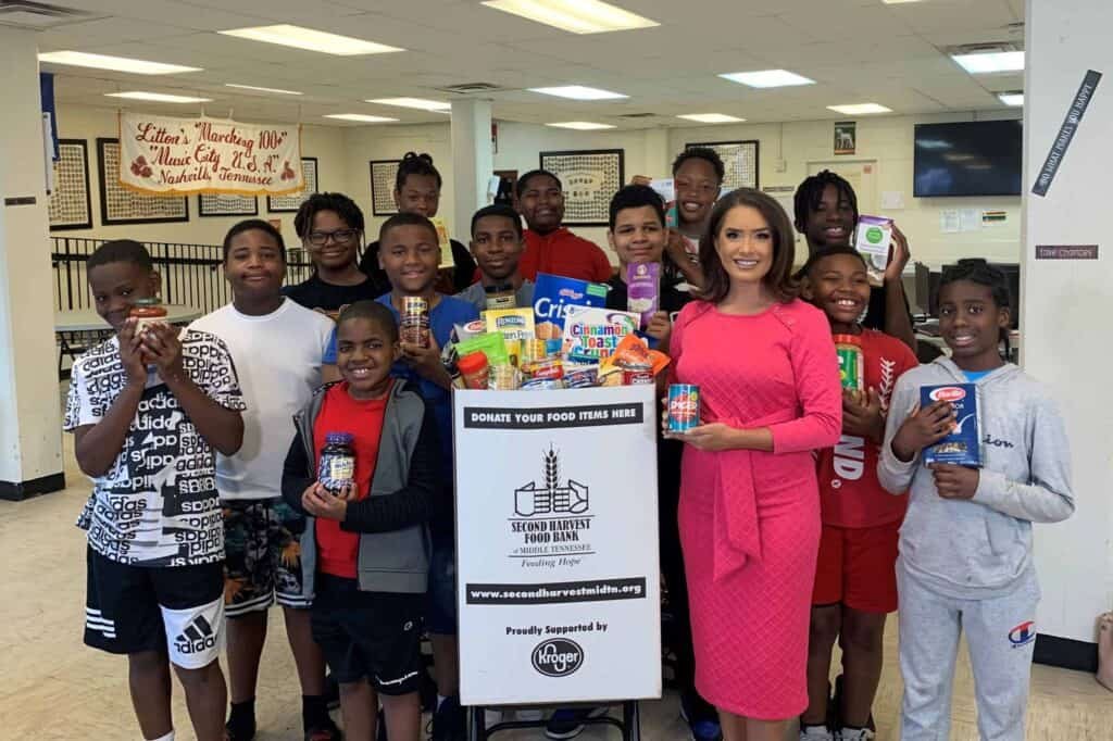 A group of smiling children and a woman in a pink dress stand around a food donation bin filled with canned goods and boxes in a school setting, supporting a Second Harvest Food Bank drive.