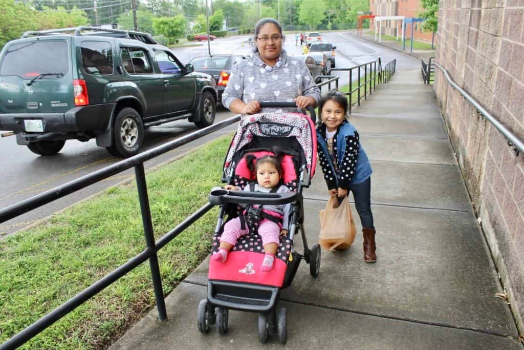 A woman pushes a stroller with a baby while a young girl holding a brown bag walks beside them on a sidewalk near parked cars and a brick wall.