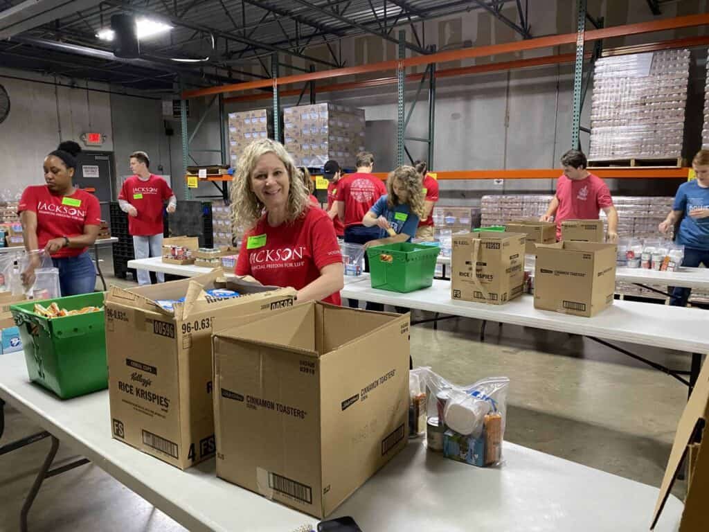 Volunteers in red shirts pack food items into boxes at tables in a warehouse, preparing supplies for distribution. Shelves filled with boxes are visible in the background.