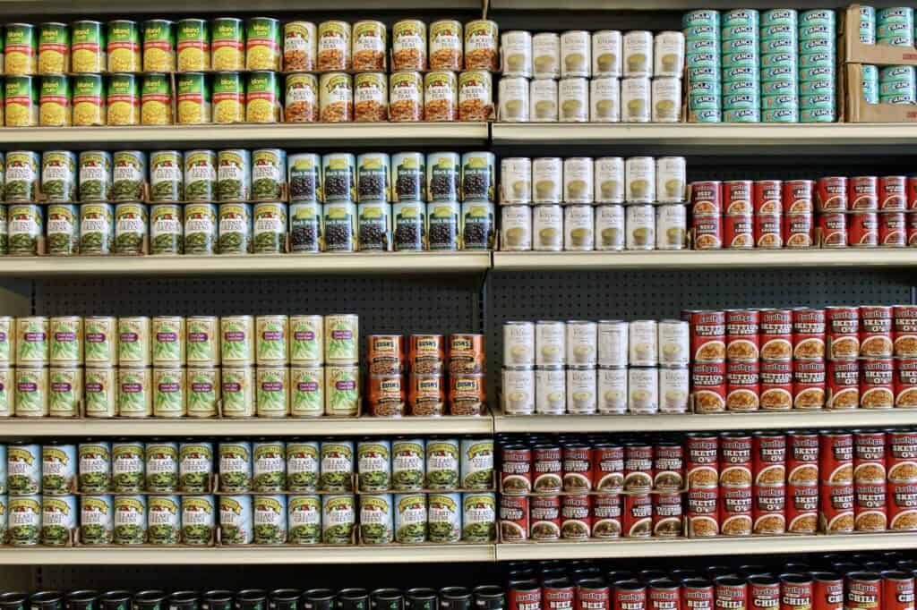 Shelves stocked with neatly arranged canned food, including vegetables, beans, soup, and tuna, in various colorful labels and sizes, organized by type and stacked in rows at a grocery store.