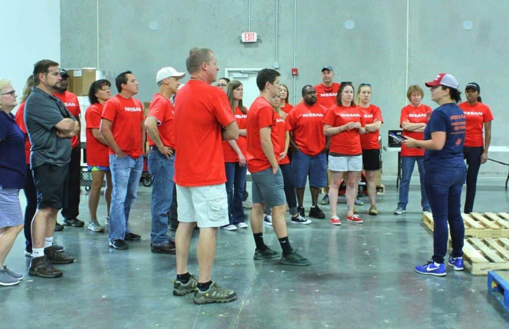 A group of people wearing matching red Nissan shirts stand in a warehouse, listening to a woman in a blue cap and jeans who appears to be giving instructions. Wooden pallets and boxes are visible in the background.