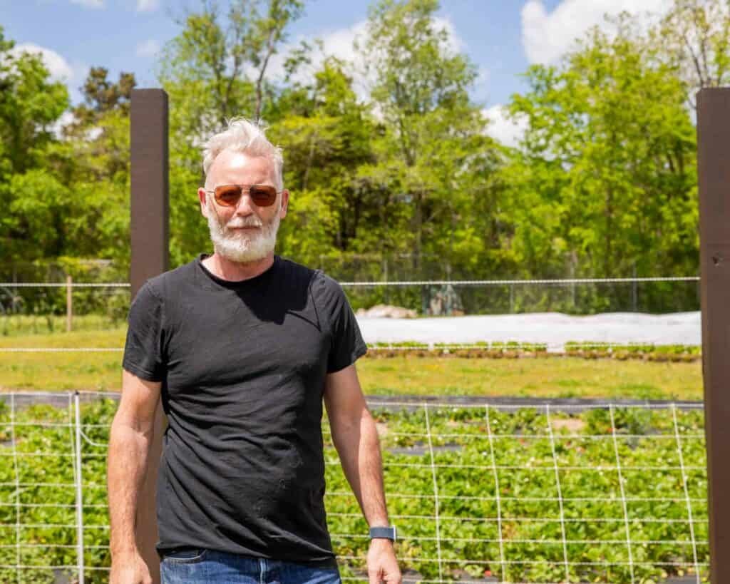 A man with white hair and a beard, wearing sunglasses, a black t-shirt, and jeans, stands outdoors in front of a wire fence and greenery on a sunny day.