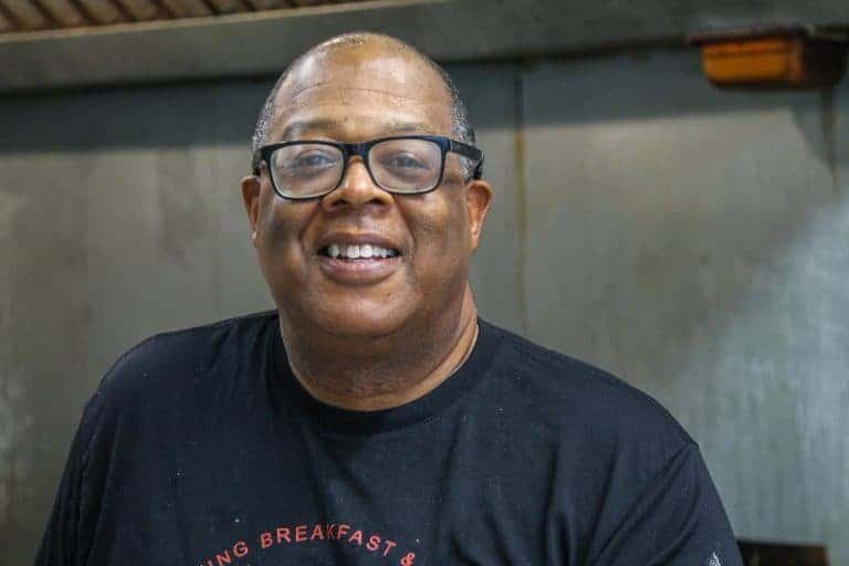 A man with glasses smiles at the camera, wearing a black T-shirt and standing in what appears to be a kitchen with a metallic background.