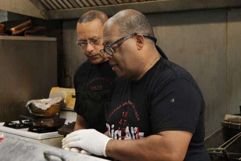 Two men wearing black shirts and aprons work together in a kitchen. One is preparing food with gloved hands while the other observes. They appear focused and are surrounded by kitchen equipment.