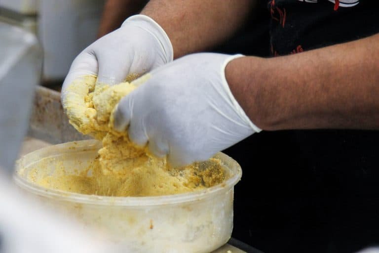 A person wearing white gloves mixes or kneads a yellow, crumbly dough or mixture in a large plastic container, suggesting food preparation in a kitchen or restaurant setting.