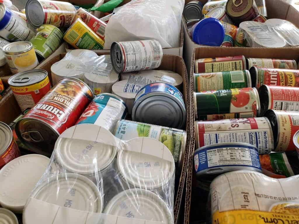 Boxes filled with assorted canned goods, including vegetables, soups, and beans, organized together, likely for donation or distribution. Some cans are wrapped in plastic, and labels are visible on many of the items.