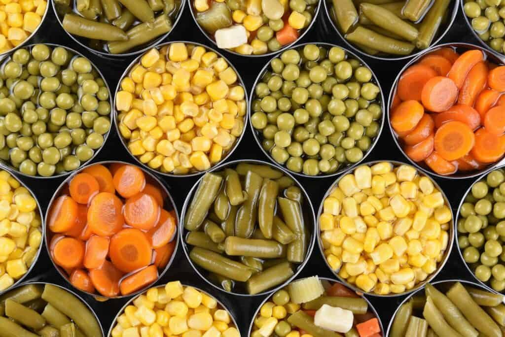 An overhead view of open cans filled with various vegetables, including corn, peas, green beans, carrots, and mixed vegetables, arranged closely together in a grid pattern.