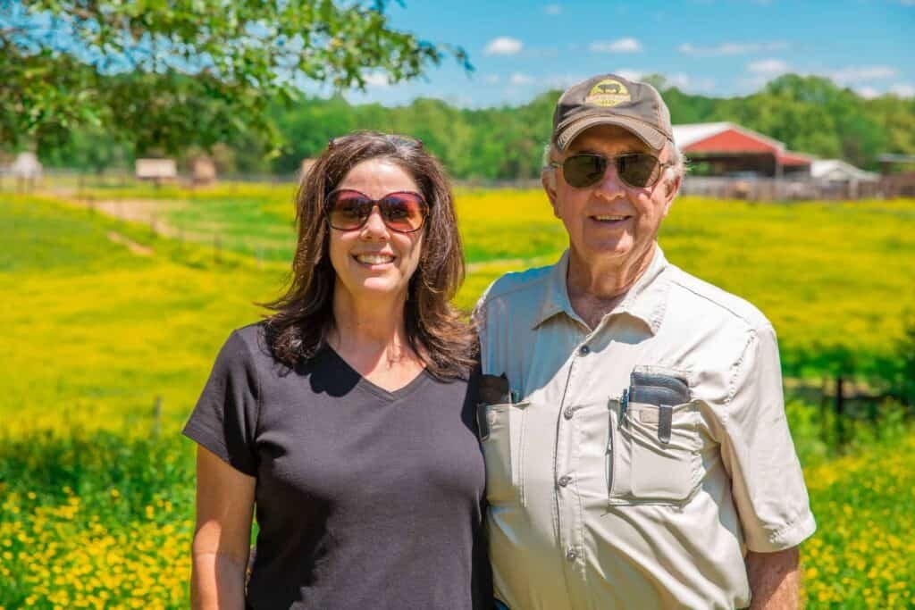 Two people wearing sunglasses stand smiling in a sunny field of yellow flowers, with barns and trees visible in the background. The scene is bright, relaxed, and outdoorsy.