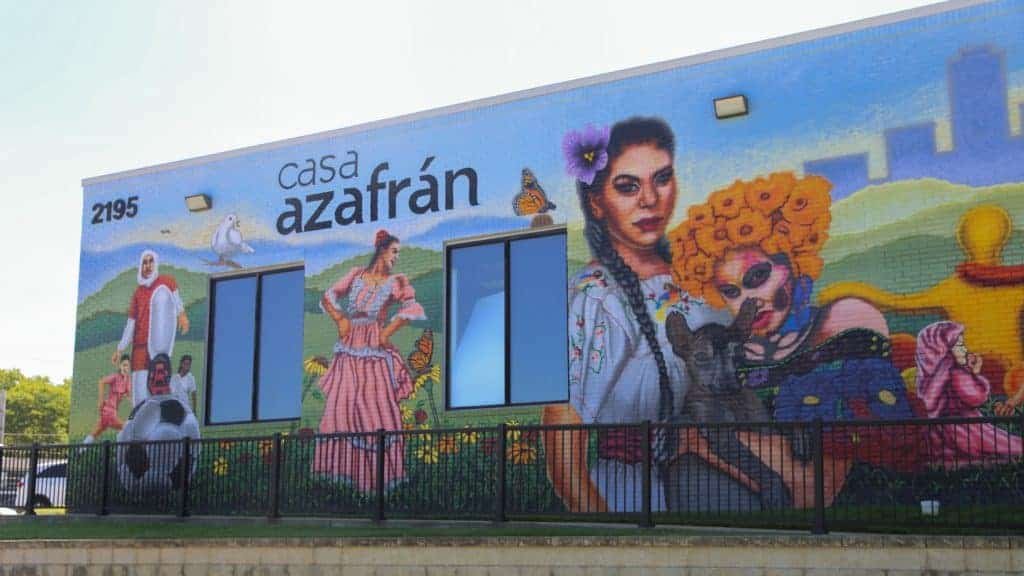A colorful mural on the Casa Azafrán building features vibrant portraits, including women in traditional dress, a child with a soccer ball, and lively patterns, celebrating Latino culture. The address 2195 is visible.