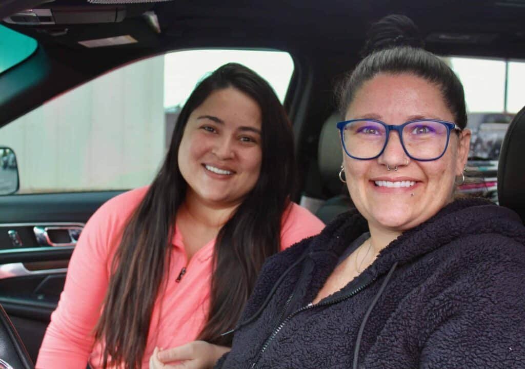 Two women smiling while sitting in the front seats of a car. One woman is wearing a pink top with long dark hair, and the other is wearing glasses and a dark jacket, both looking at the camera.
