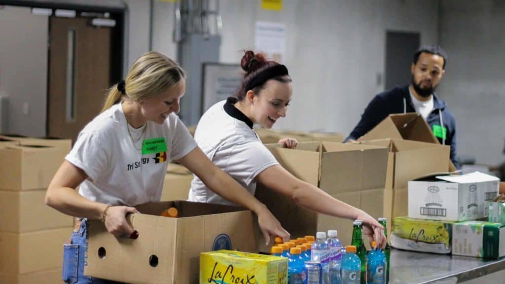 Three people pack boxes with drinks and other supplies at a table in what appears to be a warehouse. Two women smile while packing, and a man stands nearby, also assisting.