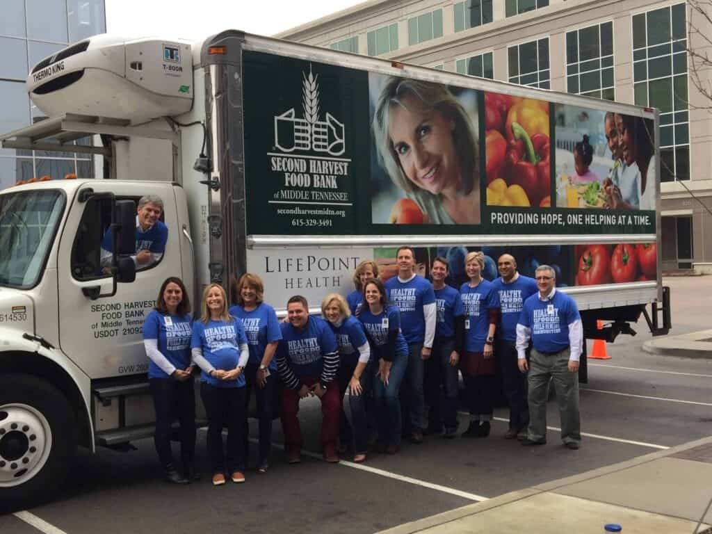 A group of people wearing matching blue Healthy Food, Healthy Future shirts stand smiling in front of a Second Harvest Food Bank truck parked near a modern building.