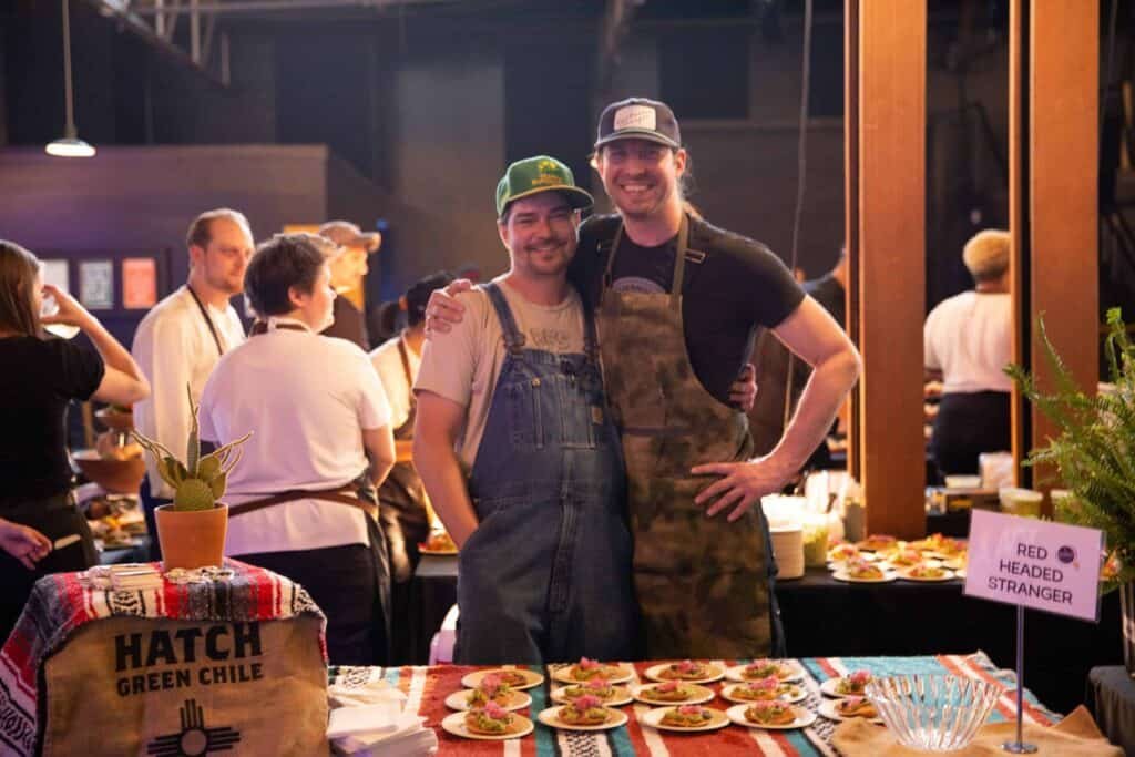 Two smiling men in aprons stand together behind a table filled with plated food at a busy indoor event. A Red Headed Stranger sign and a Hatch green chile bag are visible on the colorful tablecloth.