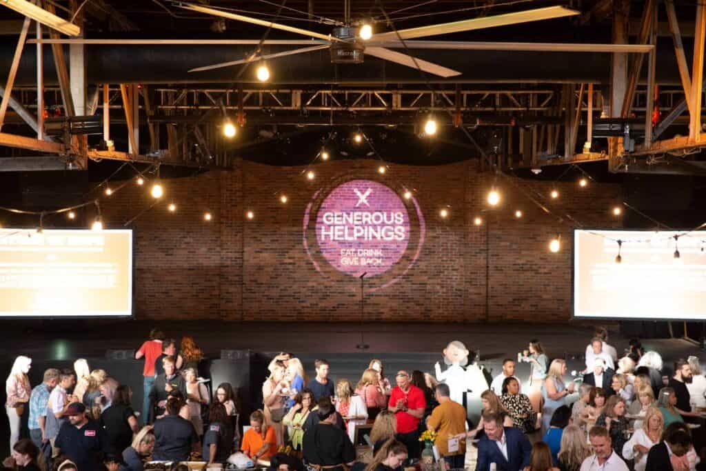 A large group of people mingles inside an industrial-style event space with exposed beams. A lit sign on the brick wall reads “Generous Helpings: Eat. Drink. Give Back.”.