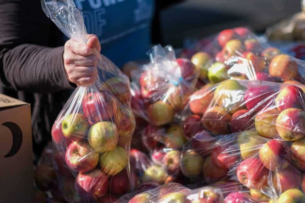 A person holds a clear plastic bag filled with apples, surrounded by more bags of apples on a table, suggesting a market or food distribution setting.