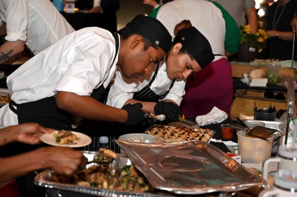 Two chefs wearing black hats and white uniforms carefully plate food at a busy event, surrounded by trays of prepared dishes, utensils, and other people in the background.