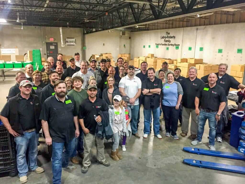 A large group of people pose and smile together inside a warehouse, surrounded by cardboard boxes and industrial shelves. Most of them are wearing casual clothes and name tags, suggesting a team event or volunteer activity.
