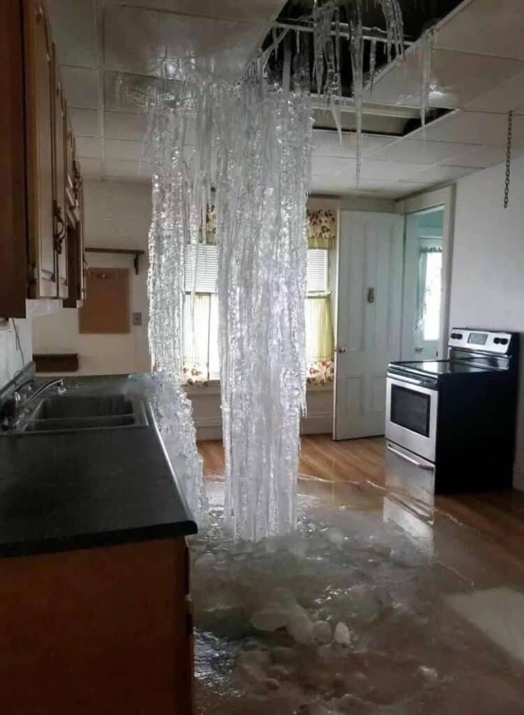 A kitchen with a large cluster of icicles hanging from and through a hole in the ceiling, with ice covering part of the floor below, suggesting severe water leak and freezing conditions.