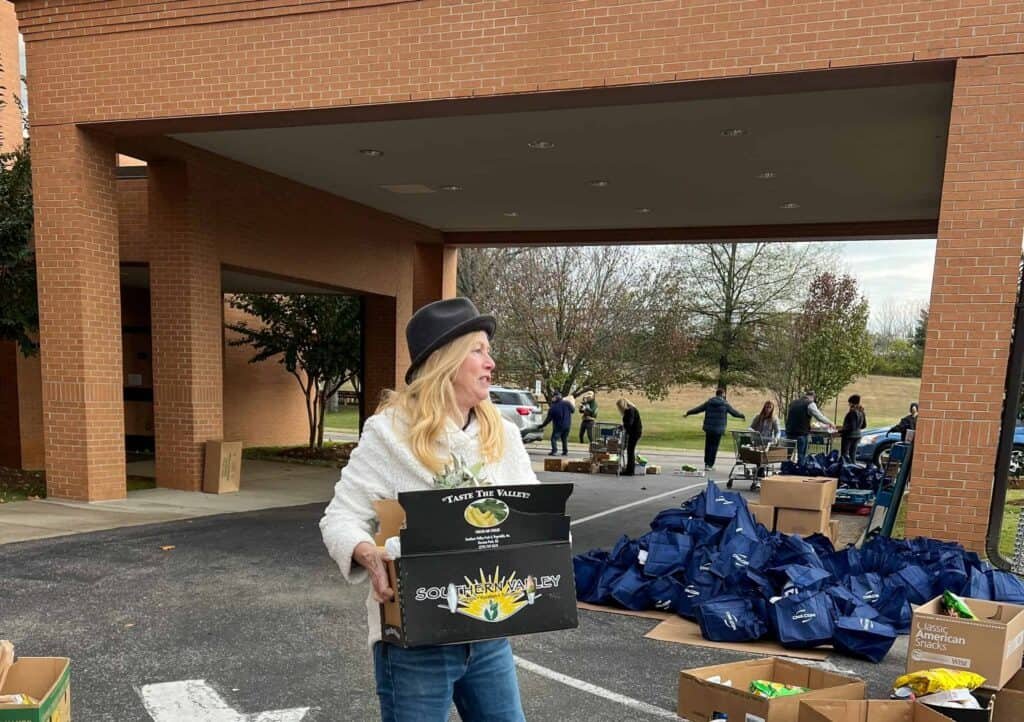 A woman in a hat and white jacket carries a box outside a building, while people in the background organize blue bags and boxes under a covered driveway. Trees and parked cars are visible in the background.