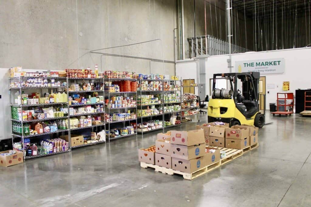 A warehouse with shelves stocked with various food items, a pallet of produce boxes in the foreground, and a yellow forklift parked nearby. A sign on the wall reads “The Market.”.