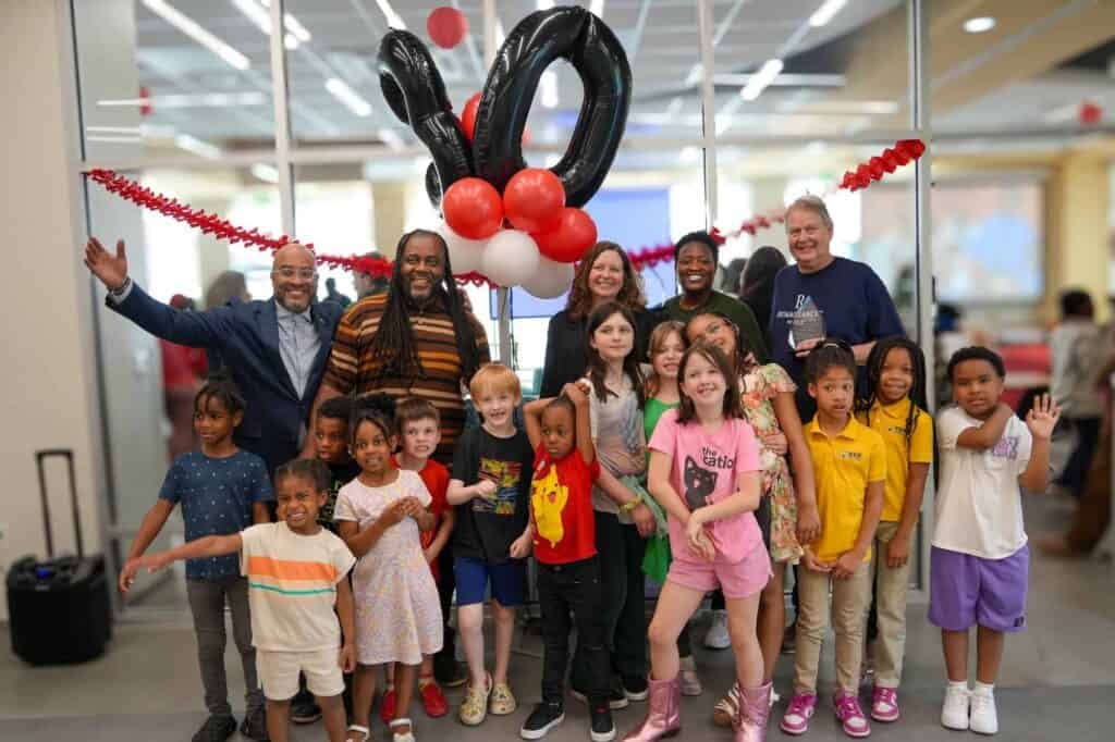 A diverse group of adults and children smile and pose together indoors at a festive event with balloons, red and white garlands, and a large 10 balloon decoration in the background.
