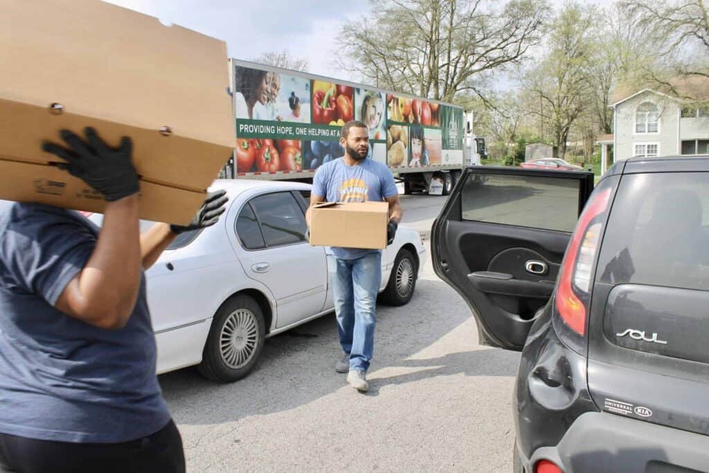 Two people load boxes into cars on a street. One person is placing a box into the back seat of a black car, while another person carries a box. A truck with a colorful produce wrap is parked in the background.