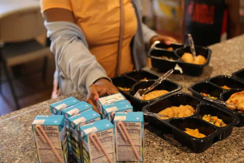 A person in a yellow shirt reaches for a meal tray on a counter with several black meal trays filled with food and multiple cartons of milk in the foreground.