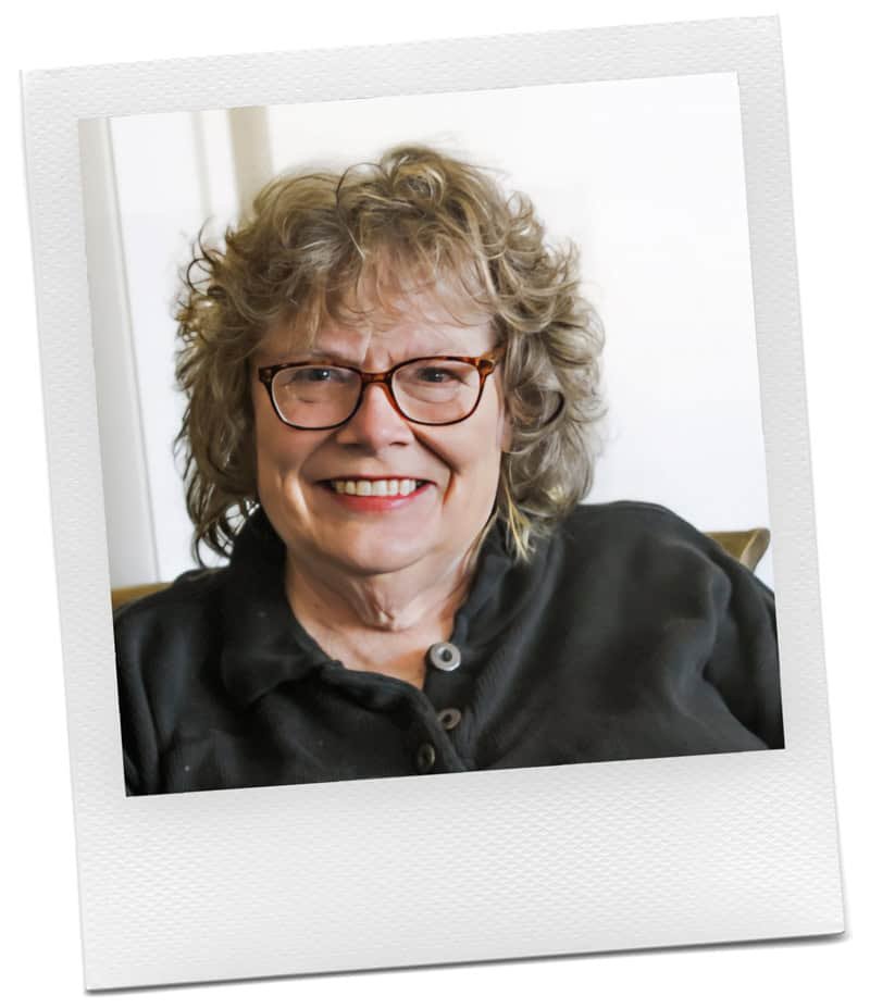 A woman with curly, light brown hair and glasses smiles at the camera. She is wearing a dark shirt and is seated indoors. The image has a white Polaroid-style border.
