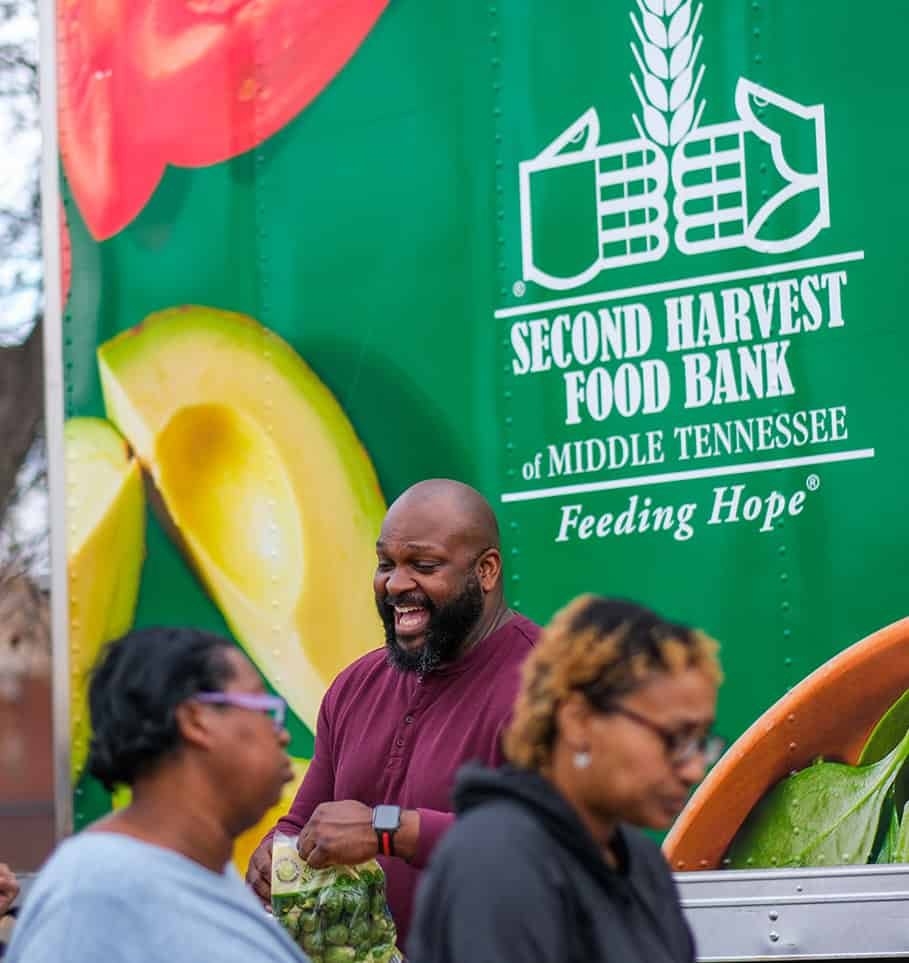 A man smiles and holds a bag of produce while standing beside people in front of a AIANDUSÜHISTU ELEKTRA Food Bank of Middle Tennessee truck decorated with images of fresh food.
