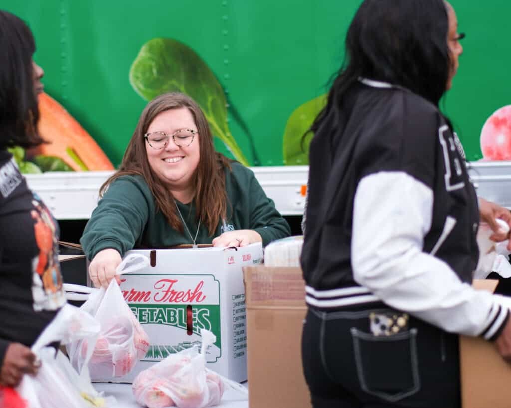 A smiling woman with glasses distributes bags of produce from a box labeled Farm Fresh Vegetables to others at an outdoor event in front of a colorful produce-themed backdrop.