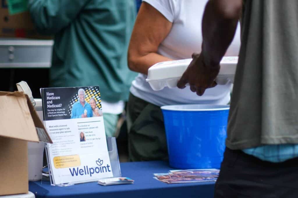 A person hands a takeout container to another person at a table covered with brochures and a Wellpoint sign, suggesting a community event or outreach. A blue cooler and informational pamphlet are also visible on the table.