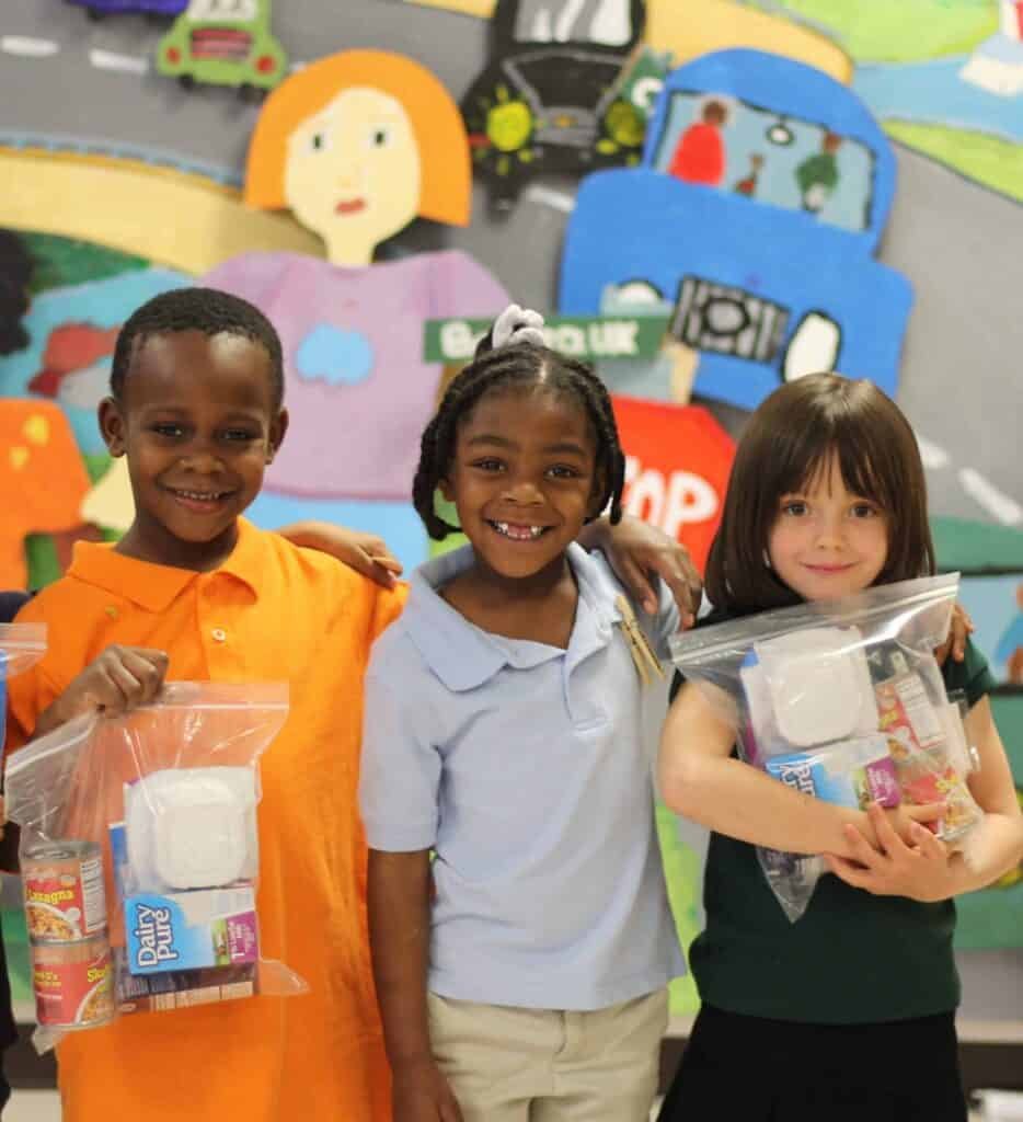 Three smiling children stand together holding clear bags with food items in front of a colorful mural featuring people, cars, and a road.