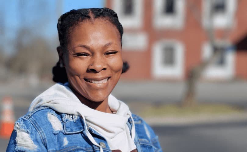 A woman with braided hair smiles outdoors, wearing a denim jacket over a light-colored hoodie. The background is blurred, showing a red brick building and trees in daylight.