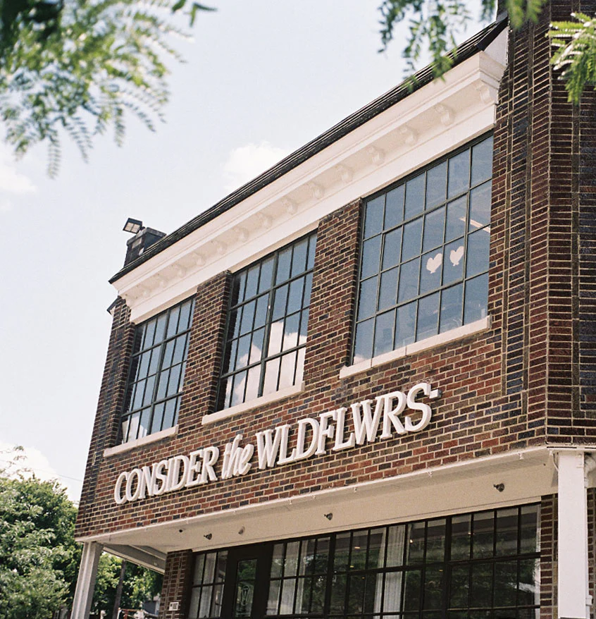 A brick building with large windows displays a sign reading CONSIDER the WLDFLWRS. Three paper hearts are taped to one window, and green tree branches frame the image.