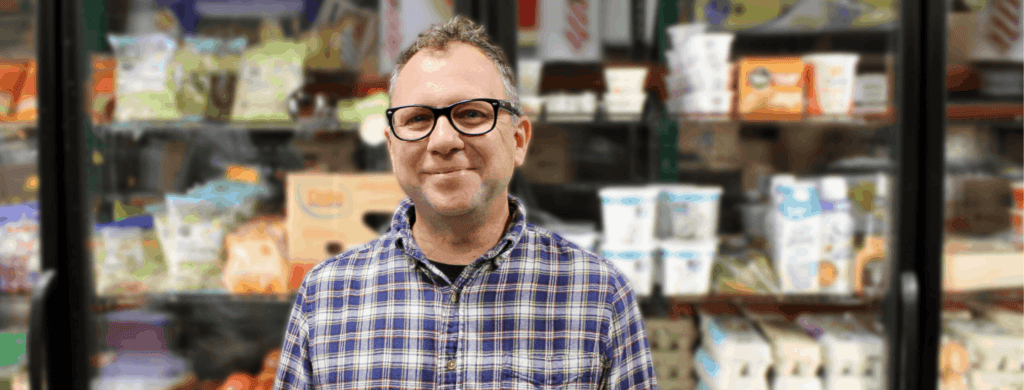 A person wearing glasses and a plaid shirt smiles while standing in front of a grocery store freezer filled with various packaged products.