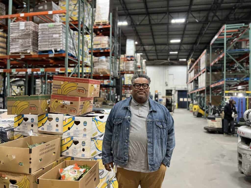 A man wearing glasses, a gray shirt, and a denim jacket stands in a warehouse filled with boxes of produce and tall shelves stacked with goods. The space is industrial with high ceilings and forklifts in the background.