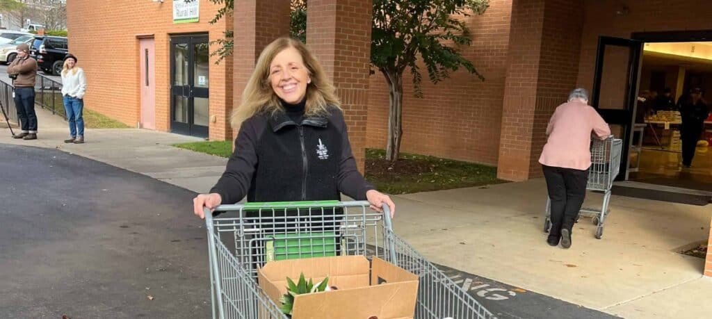 A smiling woman pushes a shopping cart with a cardboard box outside a brick building; other people with carts are visible in the background near the entrance.