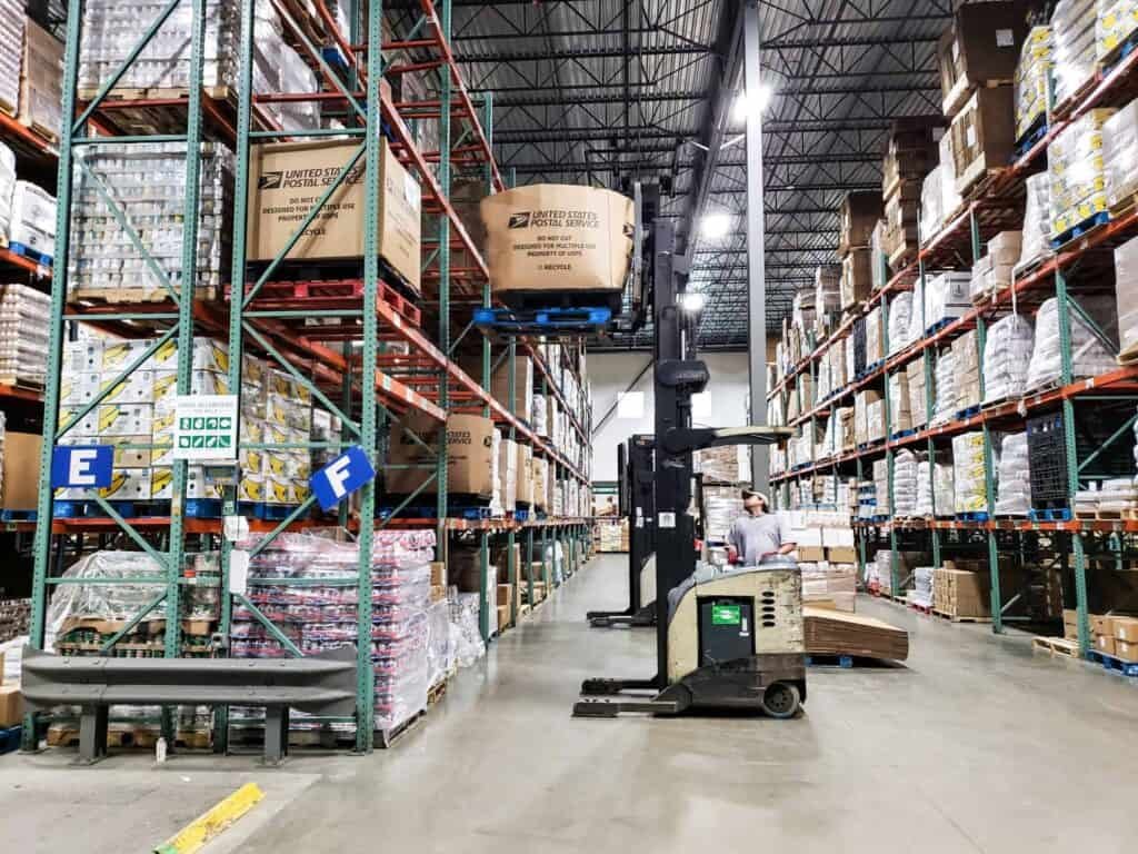 A warehouse interior with tall shelves stacked with boxes and pallets. A forklift is lifting a box, and a person is visible in the aisle. The floor is clean and the area is brightly lit.