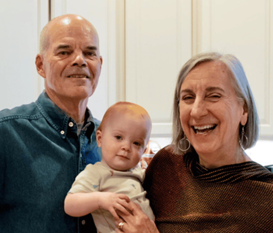 An older man and woman are smiling and standing close together in a kitchen, holding a baby between them. The woman is laughing, and the baby is looking at the camera. All three appear happy and relaxed.