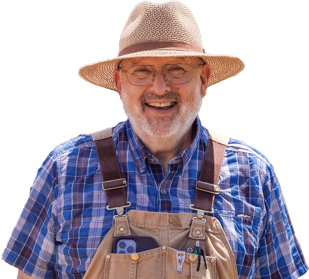 An older man with a white beard and glasses smiles, wearing a straw hat, plaid shirt, and overalls with pens and a phone in the front pocket, standing against a white background.