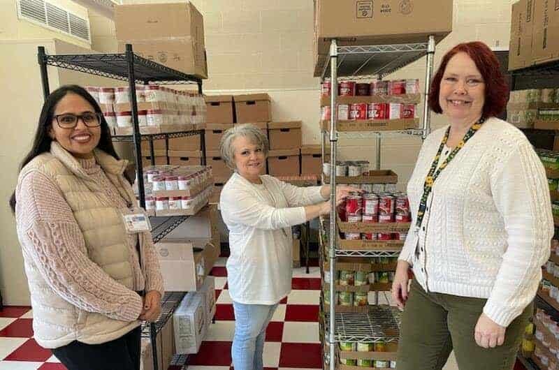 Three women stand and smile in a food pantry, surrounded by shelves stocked with canned goods and boxes. One woman organizes cans on a metal shelf while the others face the camera.