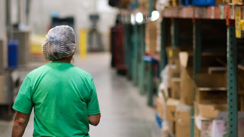 A person wearing a hair net and green shirt walks through a warehouse aisle lined with shelves of boxes and supplies. The image is focused on the persons back and the industrial setting.