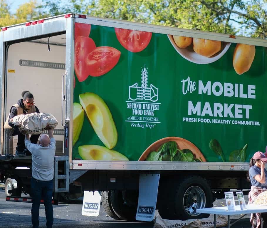 A man hands a sack of potatoes to another man standing inside a Second Harvest Food Bank Mobile Market truck, which is parked outdoors with a colorful fresh produce graphic on its side.