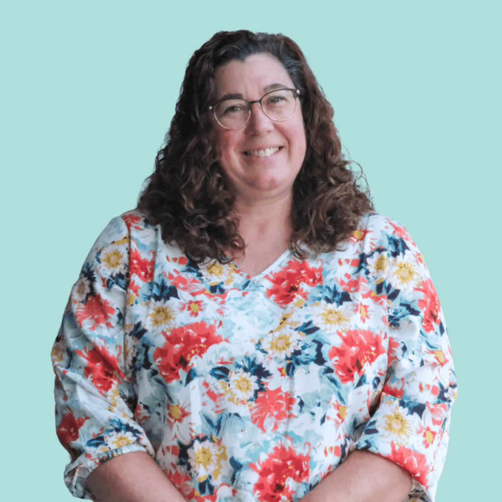 A woman with curly brown hair and glasses smiles, wearing a white top with a colorful red, yellow, and blue floral pattern, standing against a plain light teal background.