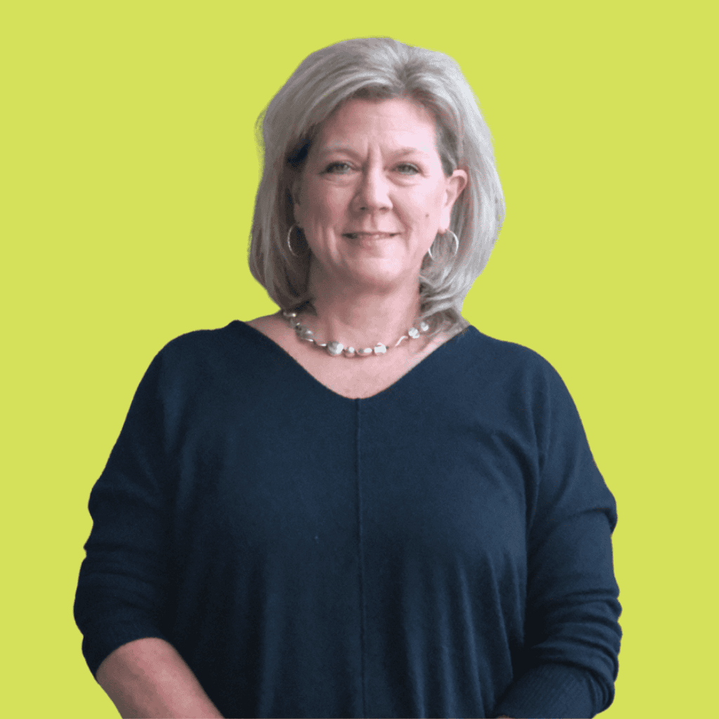 A woman with shoulder-length blonde hair, wearing a dark sweater and a beaded necklace, stands facing the camera and smiles slightly. The background is a solid light yellow-green color.