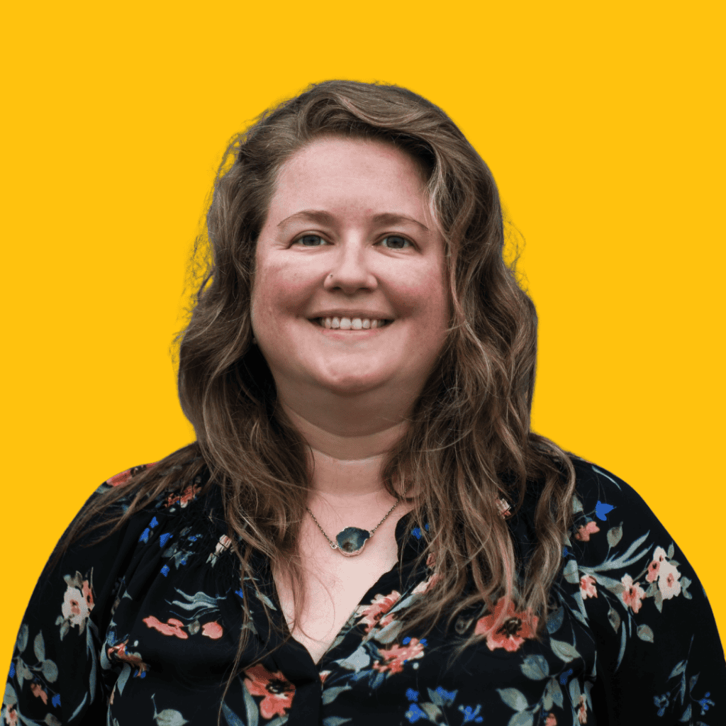 A woman with wavy brown hair, wearing a black floral top and a stone necklace, smiles in front of a bright yellow background.