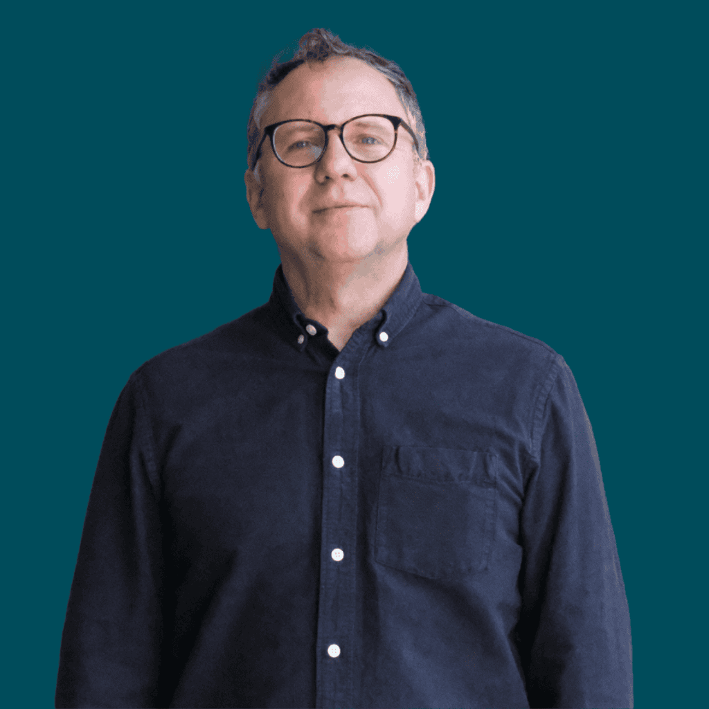A man wearing glasses and a dark button-up shirt stands in front of a solid teal background, looking at the camera with a neutral expression.