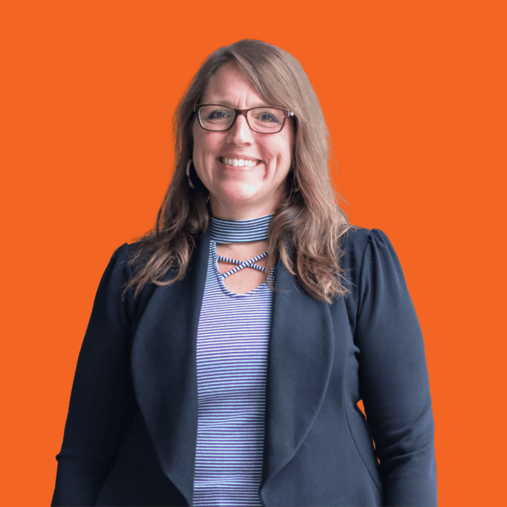 Smiling woman with long brown hair and glasses wearing a dark blazer over a striped shirt, standing in front of a bright orange background.