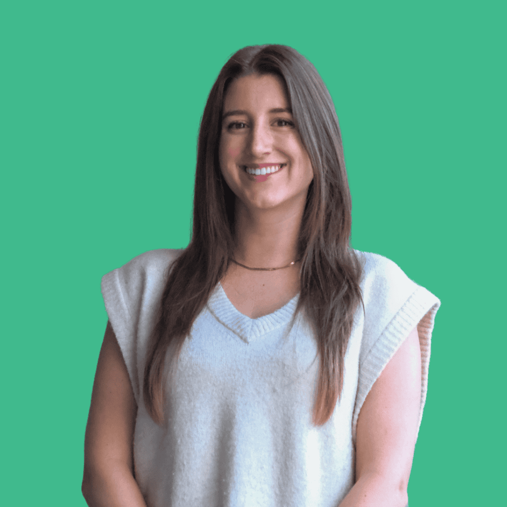 A woman with long brown hair wearing a sleeveless light gray sweater smiles while standing against a solid teal-green background.